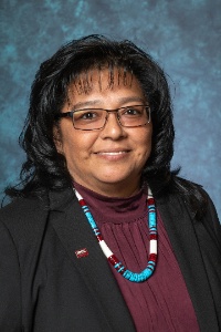 Picture of Michelle Lonasee, Program Coordinator of American Indian Program. The image is a portrait of an individual facing the camera against a blue mottled background. The person has shoulder-length dark hair with bangs and wears rectangular glasses with black frames. They are dressed in a black blazer over a maroon blouse. Around their neck is a necklace composed of red, turquoise, and white beads. On the left lapel of their blazer, there is a small pin with white text on a red background.